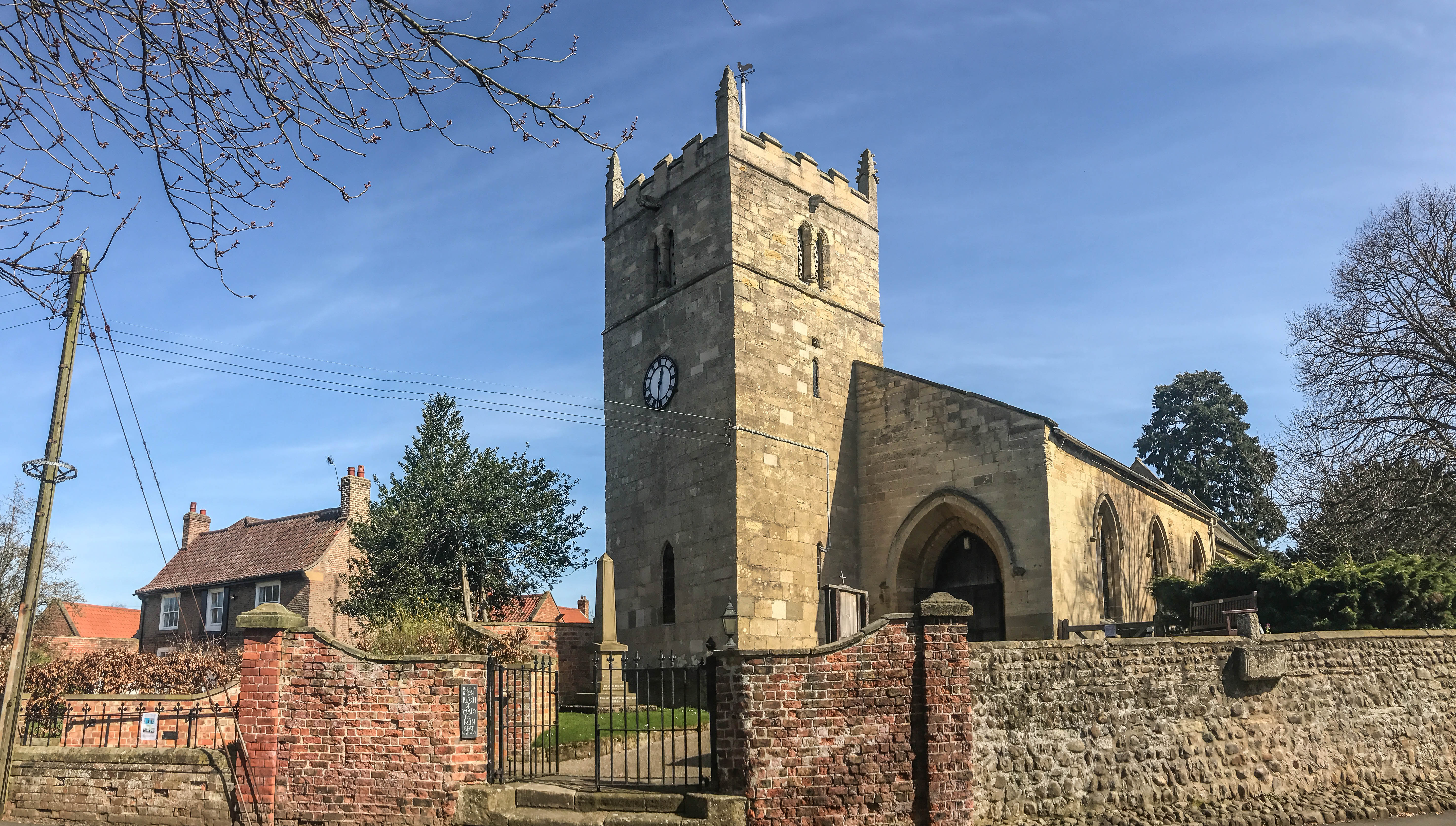 St Mary the Virgin church, Great Ouseburn — a Norman tower with clock and battlemented parapet behind a red-brick churchyard wall