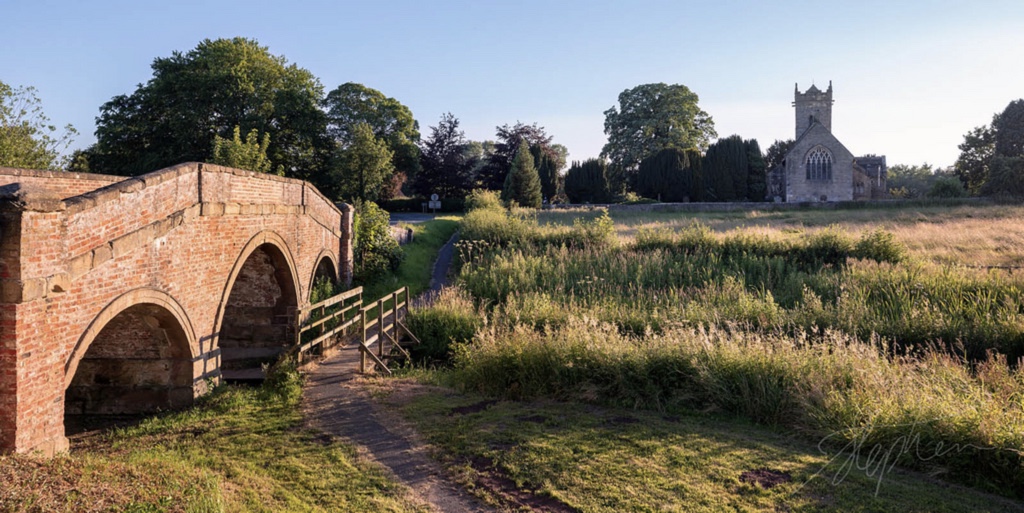 Holy Trinity church, Little Ouseburn, seen from across the meadow with the Georgian bridge in the foreground at golden hour
